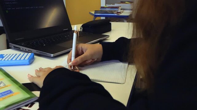 Female student writing code in a notebook while a laptop runs a Python programming example, showcasing modern education technology and artificial intelligence assistance in a classroom setting