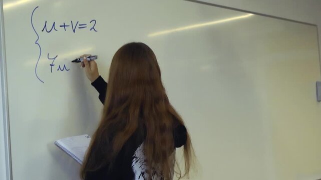 Rear view of a smart high school girl with long hair writing a system of equations on a whiteboard during a mathematics class, holding a notebook and a marker pen in her hands