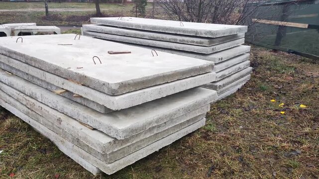 Stacks of heavy grey concrete slabs for road construction, featuring metal hooks for lifting, lying outdoors on damp ground with dry grass and sparse vegetation under overcast sky