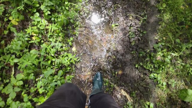 Person walking on muddy path with green wellington boots splashing in puddles of water surrounded by dense green forest foliage during daytime