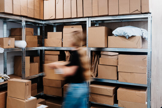 A blurred worker carries a parcel past metal shelving stacked with cardboard boxes in a small storage room. Fast order fulfillment and inventory management for a growing ecommerce business.