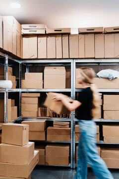 Blurred woman in motion pulls a cardboard box from metal shelving in a small storage room packed with parcels. Fast order picking and fulfillment in a growing online retail business.