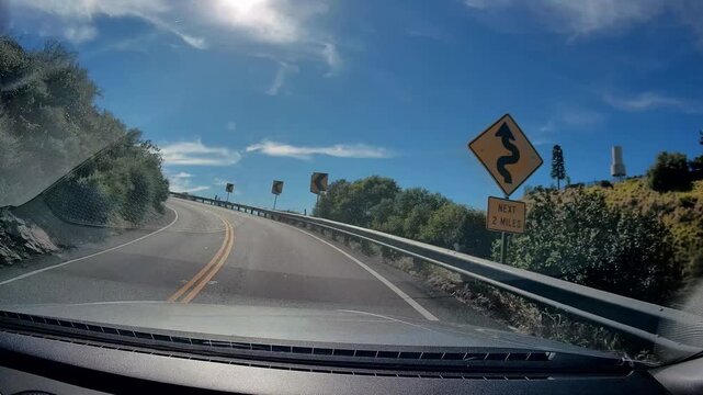Dashcam view driving on winding mountain road, sunlight on sky, green vegetation lining route, clear day conditions for travel, curve signs indicating direction of path