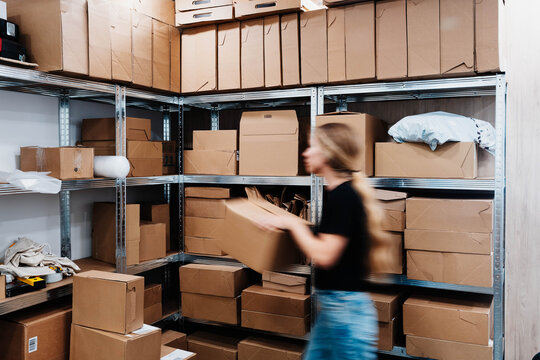 Blurred worker carries a cardboard box past metal shelves stacked with parcels in a small storage room. Order fulfillment and inventory handling in ecommerce warehouse logistics.