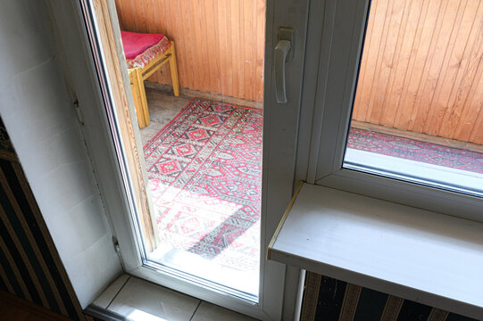 partially open white plastic balcony door reveals cozy alcove with patterned red rug, yellow stool with red cushion, warm wooden wall paneling
