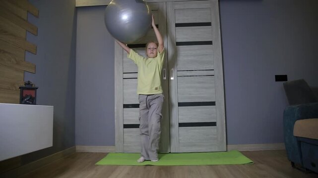 Little girl practicing fitness with exercise ball at home. Child performing physical activity on yoga mat indoors. Young female engaging in health and wellness workout.