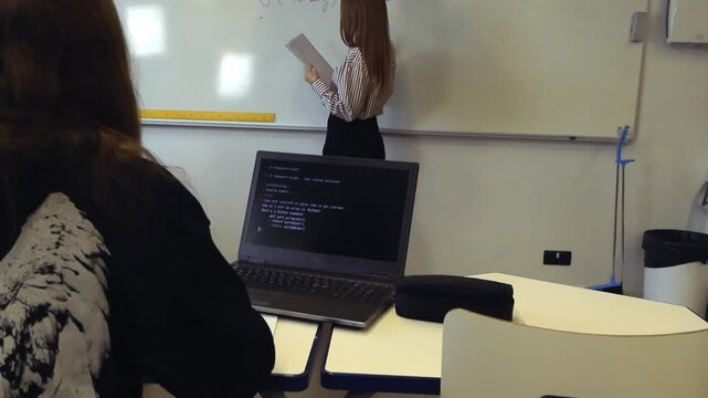 Mathematics teacher explaining a complex algebraic equation on a whiteboard during a lesson as a student with a laptop watches, showcasing the integration of technology and education
