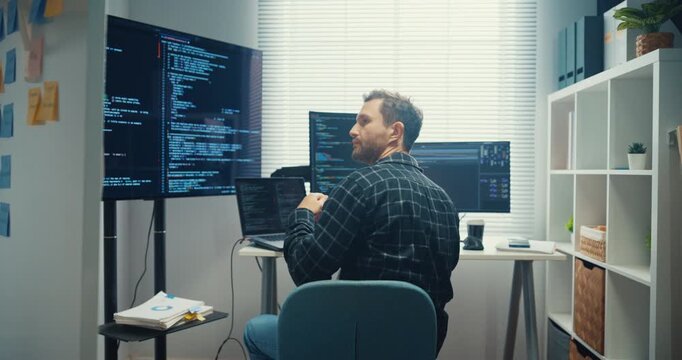 Young Caucasian male programmer writes code using multiple screens in home office workspace. AI development, cybersecurity, network database system, software engineering, remote tech startup concept.
