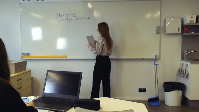 Young female teacher standing at a whiteboard, writing a system of linear equations and explaining the math problem to students during a lesson, with one pupil using a laptop in the foreground