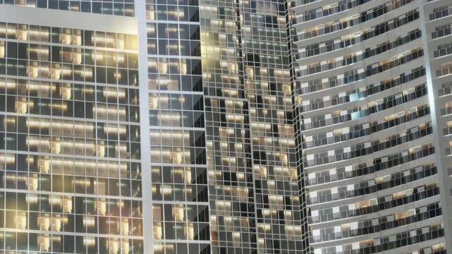 Bright windows in tall buildings are visible at night. The lights create a warm glow against the dark sky. The scene shows a busy city with many people inside.