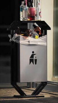 Outdoor public waste container overflowing contents with empty glass, plastic soda bottles, and discarded beer bottles, rubbish collection failure during night hours outside commercial building