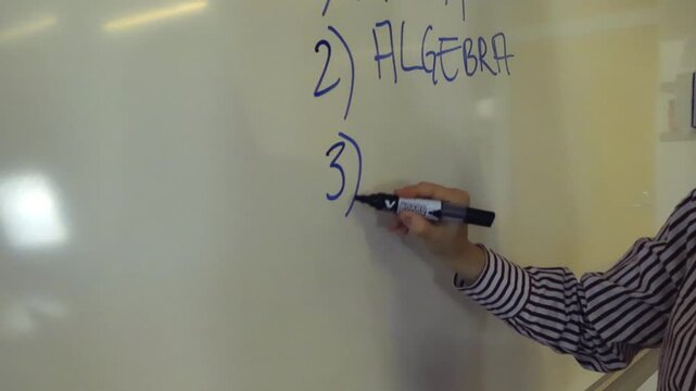 Female teacher's hand writing a numbered list of school subjects on a whiteboard during a class, listing algebra and history for her students to learn in a slow motion close up shot