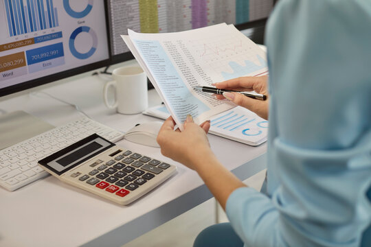 Businesswoman analyst reviewing finance report with calculator. At dual monitors she analyzes accounting data and carefully confirms monthly totals. Focused finance and analysis concept