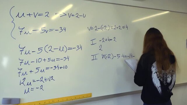 Young long haired student in a classroom writing a complex math formula on a white board with a marker, checking her notes in a notebook while solving a system of linear equations