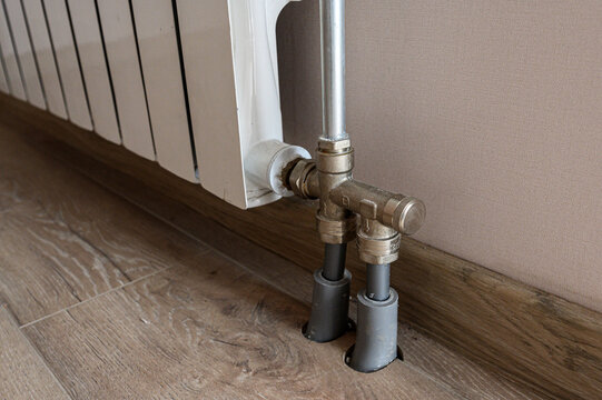 white panel radiator features bottom connection with brass fittings and shut-off valves. grey pipe sleeves protrude from floor, set against beige wall and wood-look flooring