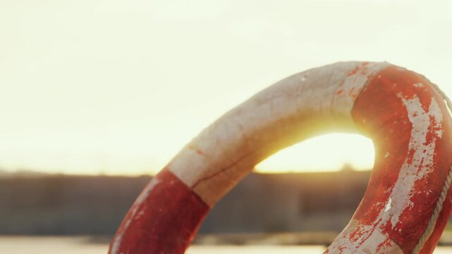 A lifebuoy rests on a surface close to the water as the sun sets in the background. Soft colors fill the sky. The scene is calm with no people present.