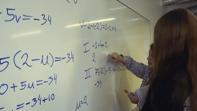 Female professor teaching algebra and pointing at a formula on the whiteboard while a student listens attentively during a lesson, highlighting the concept of education and problem solving