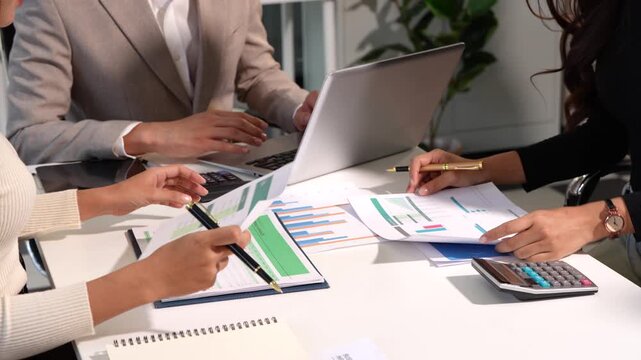 Business team reviewing printed charts, documents and a laptop at a meeting table, with a calculator and hands taking notes in a modern office.