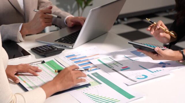 Business team reviews financial charts, reports and calculations around a table with laptop, calculators and pens during an office meeting.