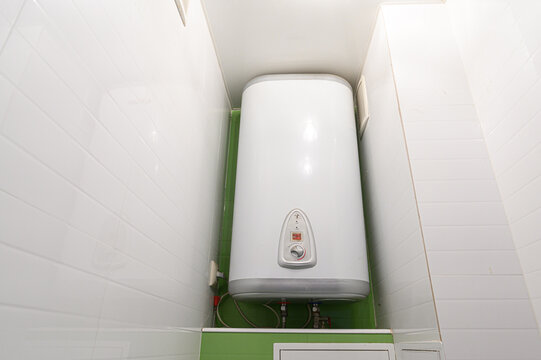 Vertical electric storage water heater in a compact tiled alcove. sleek white with control panel, pipes, and fittings at base