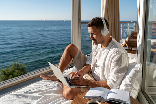 Man relaxes on bed with laptop and headphones. Ocean view through large window behind him. Sunlight bathes the room gently. He reads and works in calm, peaceful mood
