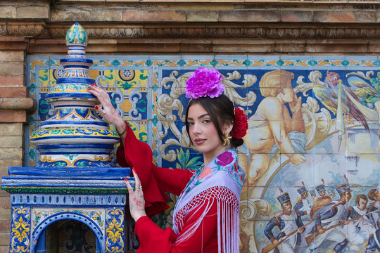 A beautiful flamenco dancer sitting on a tiled bench in a square in Seville, Spain. She is wearing a red ruffled dress and has a flower in her hair. Flamenco, a UNESCO Intangible Cultural Heritage.