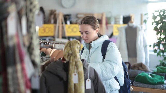 Glad adolescent female customer holding hangers with short coats during sale in retail outlet 