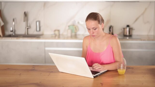 Teenage girl is sitting at a table against the background of a light kitchen and doing her homework on a laptop. Teenager participates in a video conference and listens to an online lesson. High