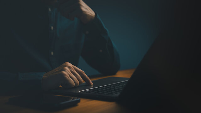 Close up of professional man hand working late at night on laptop computer technology. Focused person touching trackpad for business internet networking and online typing in dark room.