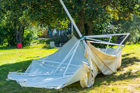 Collapsed and damaged white event tent with a bent metal frame lying broken on the green grass under a tree on a sunny day after a strong windstorm or gale.