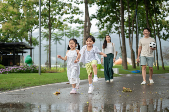 Asian family walking together through the park after rain with children running ahead on wet pathway