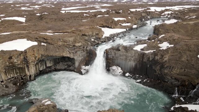Dynamic footage of powerful waterfall cascading into turquoise pool surrounded by volcanic rock formations and snow patches.