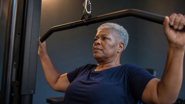 Mature African American woman performing lat pulldown exercise in gym. Strength training, healthy aging, fitness, wellness, and active lifestyle concept.