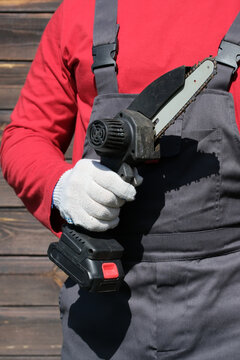 Adult man wearing work coveralls and holding a cordless electric chainsaw while working outdoors in a garden. Concept of gardening, tree maintenance and manual labor.