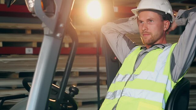 Forklift operator in safety gear relaxes in the drivers seat with hands behind his head during a work break inside an industrial warehouse.