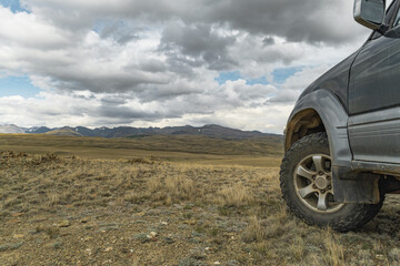 Close up of an off-road 4x4 suv car wheel with mud tire parked on a grassy hill overlooking a vast mountain valley under a dramatic cloudy sky, overlanding and road trip concept © ANDREY