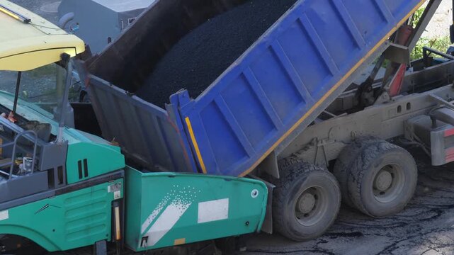 A blue dump truck is unloading hot asphalt into the hopper of a paving machine at a construction site. Heavy road equipment is performing repairs and laying new pavement.