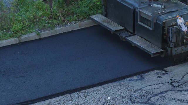 An asphalt paver smoothly spreads and lays fresh black road surface along the curb. Heavy road construction equipment performs the process of resurfacing and repairing the pavement.