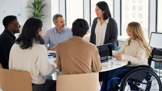 Group of diverse professionals engaged in discussion around a round table in a modern office setting with large windows and greenery in the background