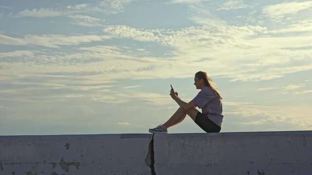Serene young woman relaxing alone outdoors, sitting on a concrete parapet against a cloudy sky, casually browsing and scrolling on her mobile phone during a peaceful, sunny day