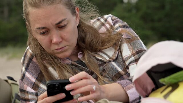 Young caucasian woman wearing a plaid shirt and laying down on a sandy beach while attentively typing and swiping on her smartphone, enjoying a moment of relaxation during a solo trip