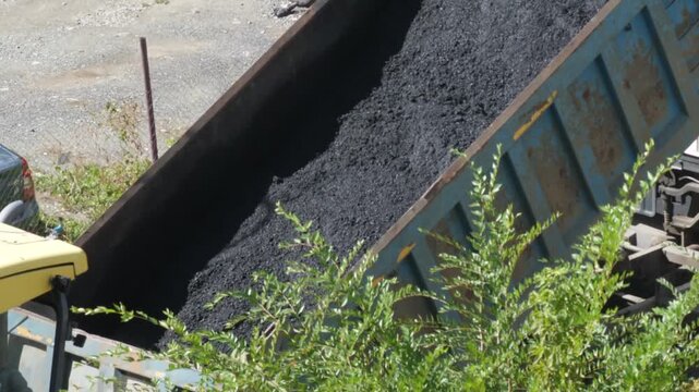 Heavy dump truck unloads a pile of hot asphalt mixture into the paver hopper during road construction works. Professional construction machinery performs paving operations on a sunny day.