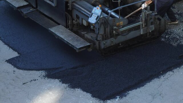 An asphalt paver machine lays a layer of fresh hot dark asphalt onto a road surface during highway construction work. 