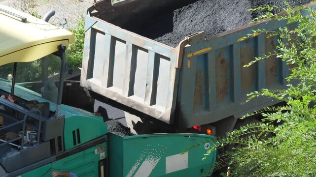 Dump truck unloading hot asphalt into the hopper of an asphalt paver during road construction works. Heavy machinery working on road repair and urban infrastructure renovation project.