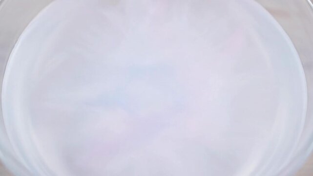 A close up view of white milk swirling into clear water in a glass bowl.