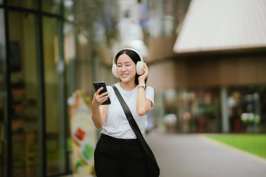 Happy Asian woman wearing headphones and checking her smartphone while walking outdoors, representing music, technology, leisure, lifestyle, and modern urban life.