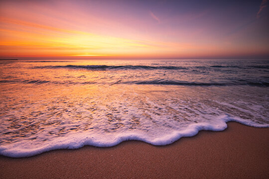 Gentle ocean wave foam washing onto sandy shore under colorful purple and orange sunset sky