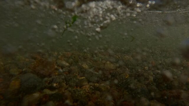 Powerful sea waves with foam and spray rise and break against the pebble shore forming many air bubbles under water surface and mixing stones on seabed, Underwater view, Slow motion