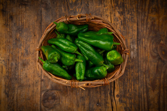 A basket holds fresh green Padron peppers on a dark wooden table