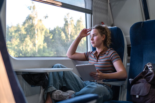 Woman sits in a train holding a tablet and looking out the window